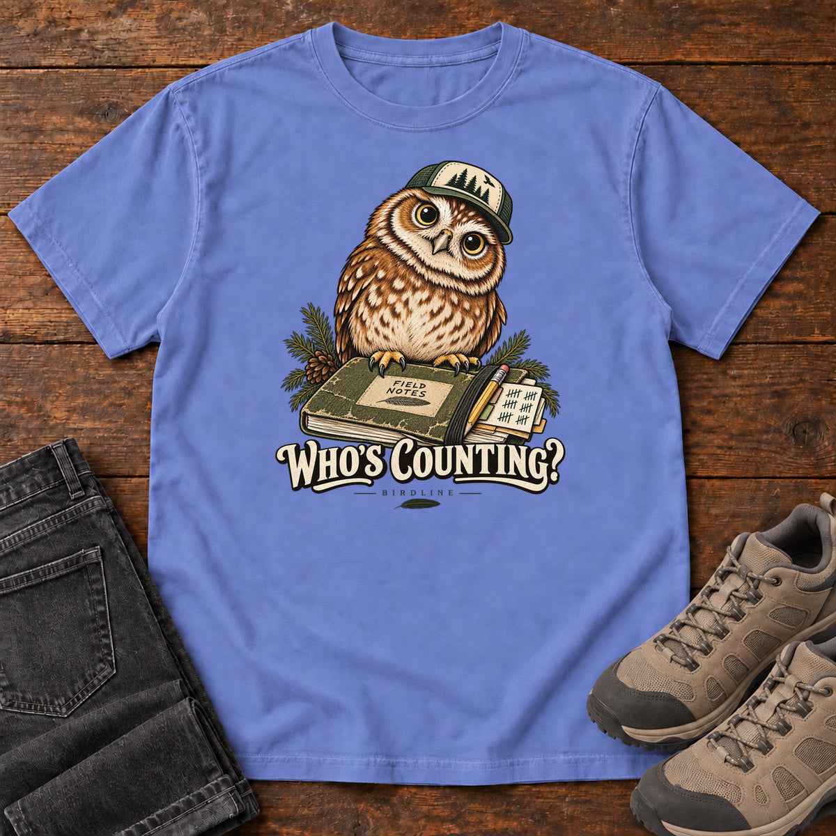 Flo Blue Who's Counting? T-Shirt with a brown owl wearing a green cap beside a field notebook and checklist above cream lettering, front flat lay on wood.