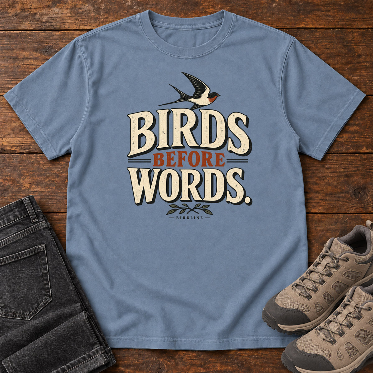 Birds Before Words T-Shirt in Blue Jean, front laid flat on wood, featuring cream and rust slogan lettering with a small flying bird above and leafy sprigs below.