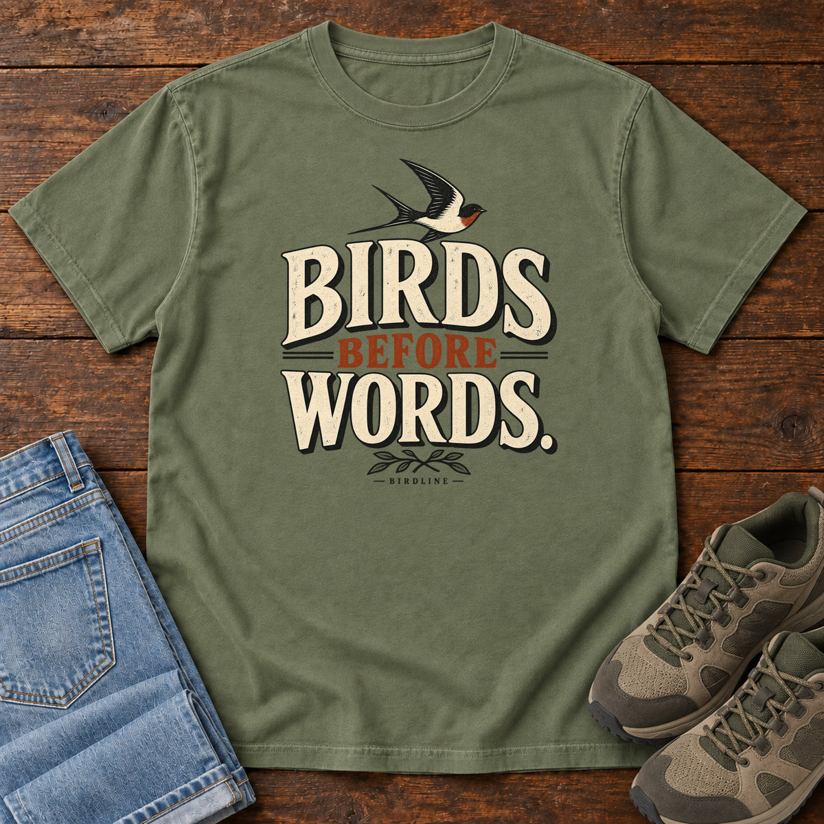 Birds Before Words T-Shirt in Moss, front laid flat on wood, featuring cream and rust slogan lettering with a small flying bird above and leafy sprigs below.