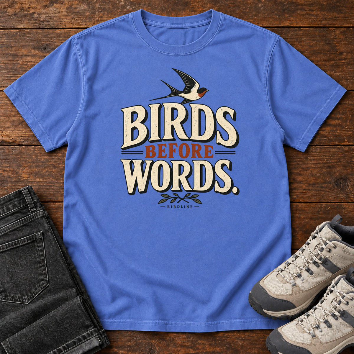 Birds Before Words T-Shirt in Mystic Blue, front laid flat on wood, featuring cream and rust slogan lettering with a small flying bird above and leafy sprigs below.