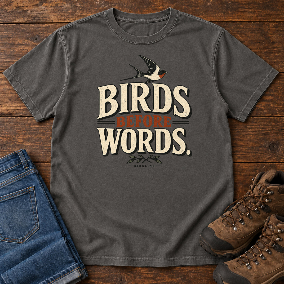 Birds Before Words T-Shirt in Pepper, front laid flat on wood, featuring cream and rust slogan lettering with a small flying bird above and leafy sprigs below.