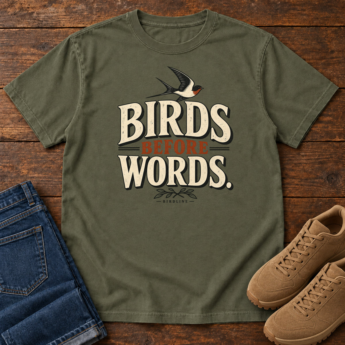 Birds Before Words T-Shirt in Sage, front laid flat on wood, featuring cream and rust slogan lettering with a small flying bird above and leafy sprigs below.