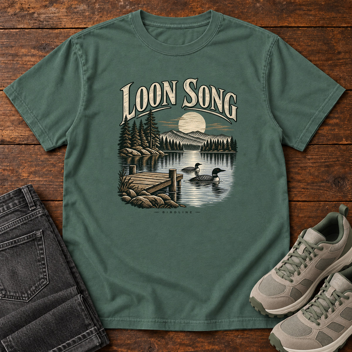 Blue Spruce Loon Song T-Shirt with two waterbirds beside a dock on a moonlit lake beneath cream lettering, front flat lay on wood.