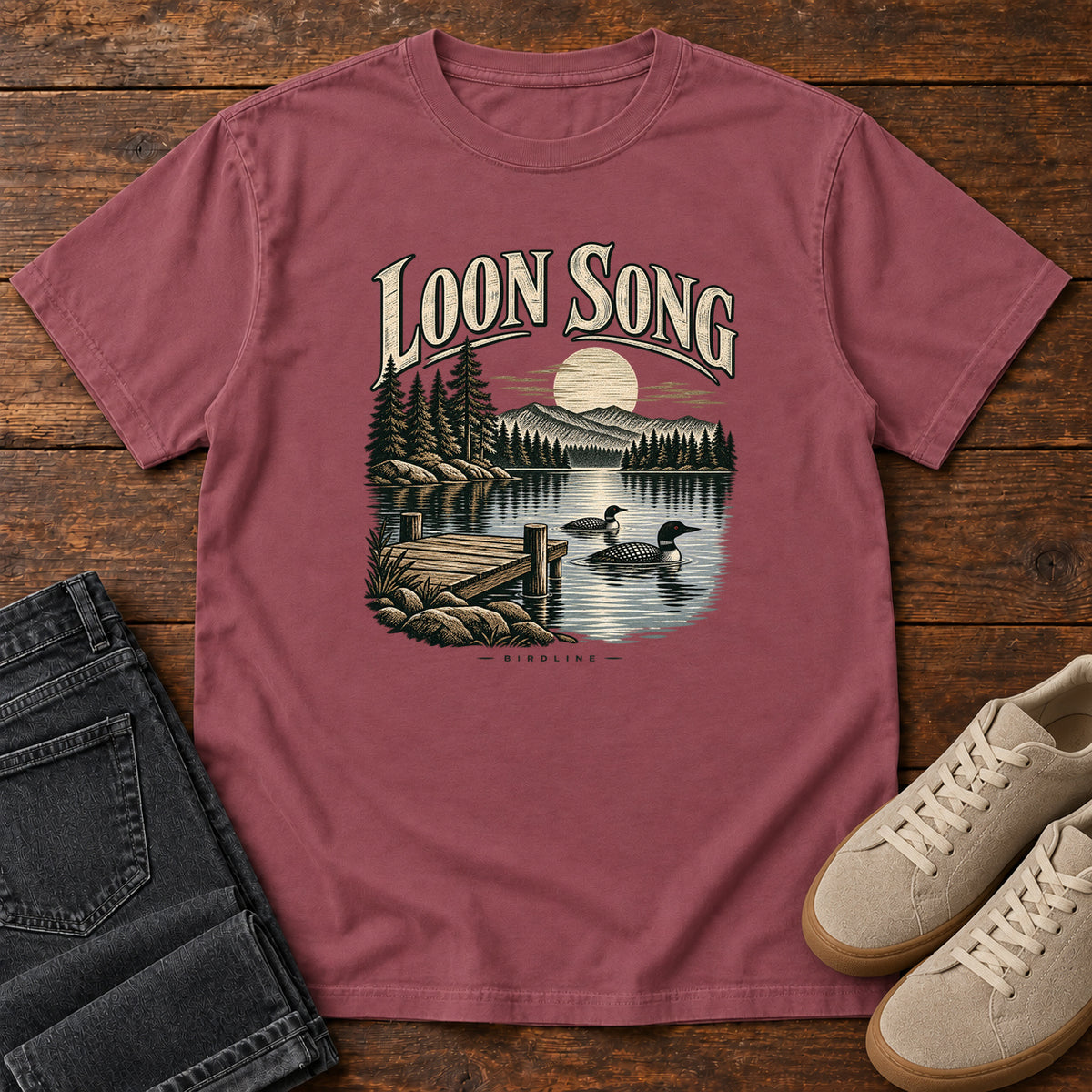 Brick Loon Song T-Shirt with two waterbirds beside a dock on a moonlit lake beneath cream lettering, front flat lay on wood.