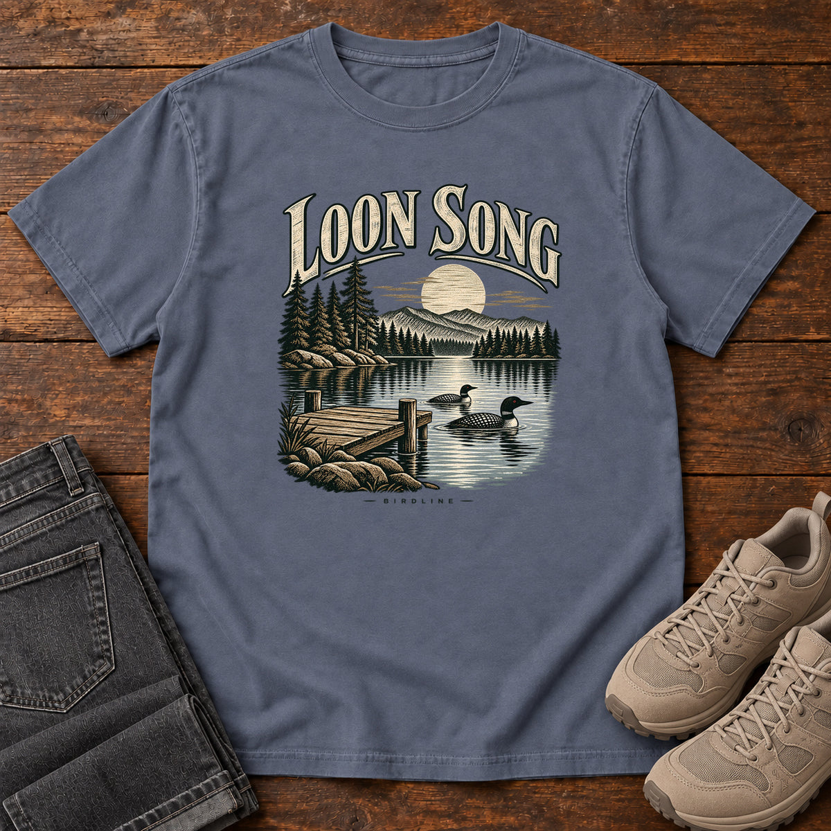 Denim Loon Song T-Shirt with two waterbirds beside a dock on a moonlit lake beneath cream lettering, front flat lay on wood.