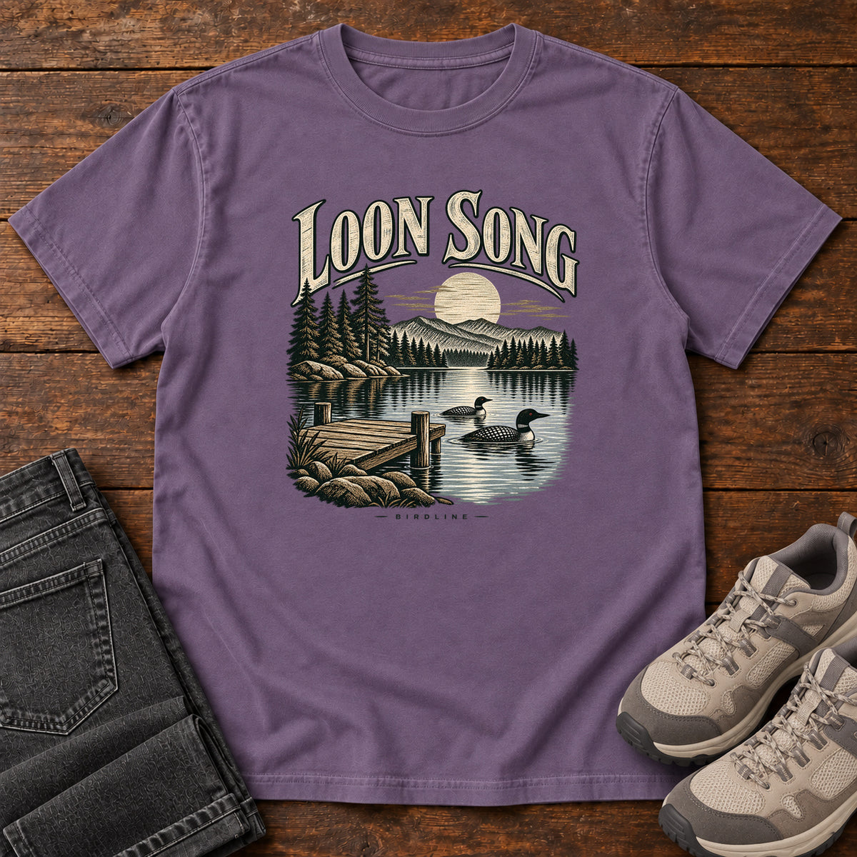 Grape Loon Song T-Shirt with two waterbirds beside a dock on a moonlit lake beneath cream lettering, front flat lay on wood.
