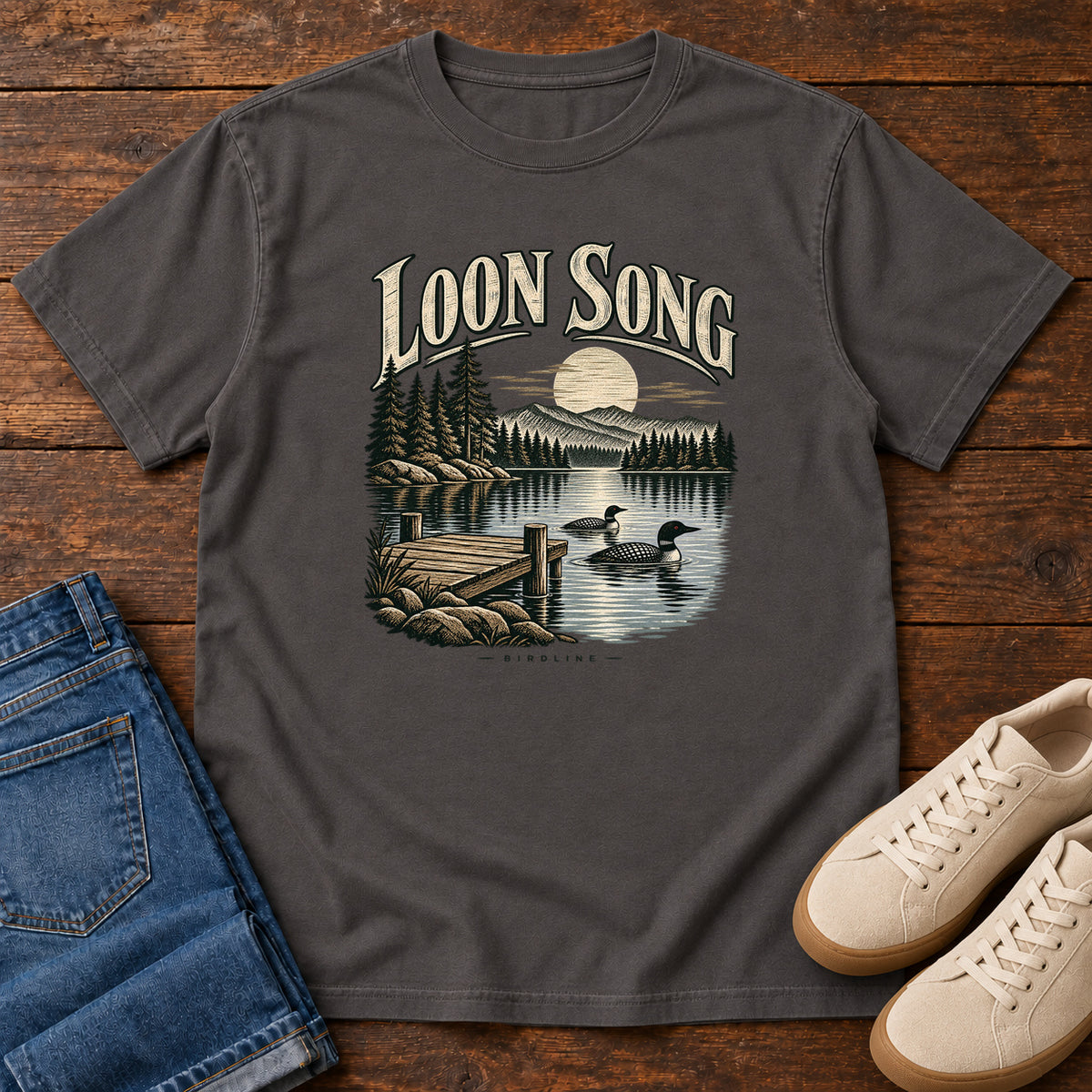 Graphite Loon Song T-Shirt with two waterbirds beside a dock on a moonlit lake beneath cream lettering, front flat lay on wood.