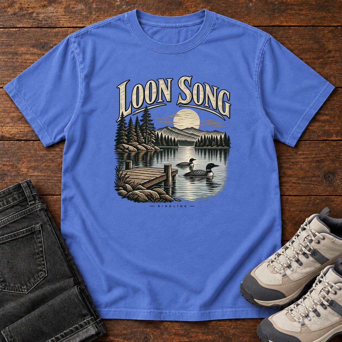 Mystic Blue Loon Song T-Shirt with two waterbirds beside a dock on a moonlit lake beneath cream lettering, front flat lay on wood.