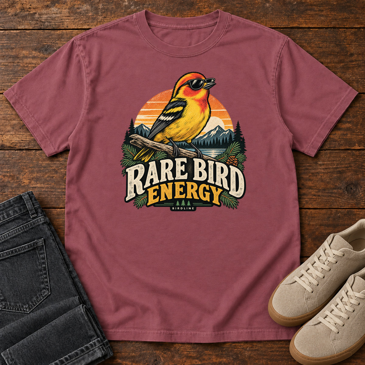 Brick Rare Bird Energy T-Shirt with a yellow and orange bird wearing sunglasses before a striped sunset; front flat lay on wood.