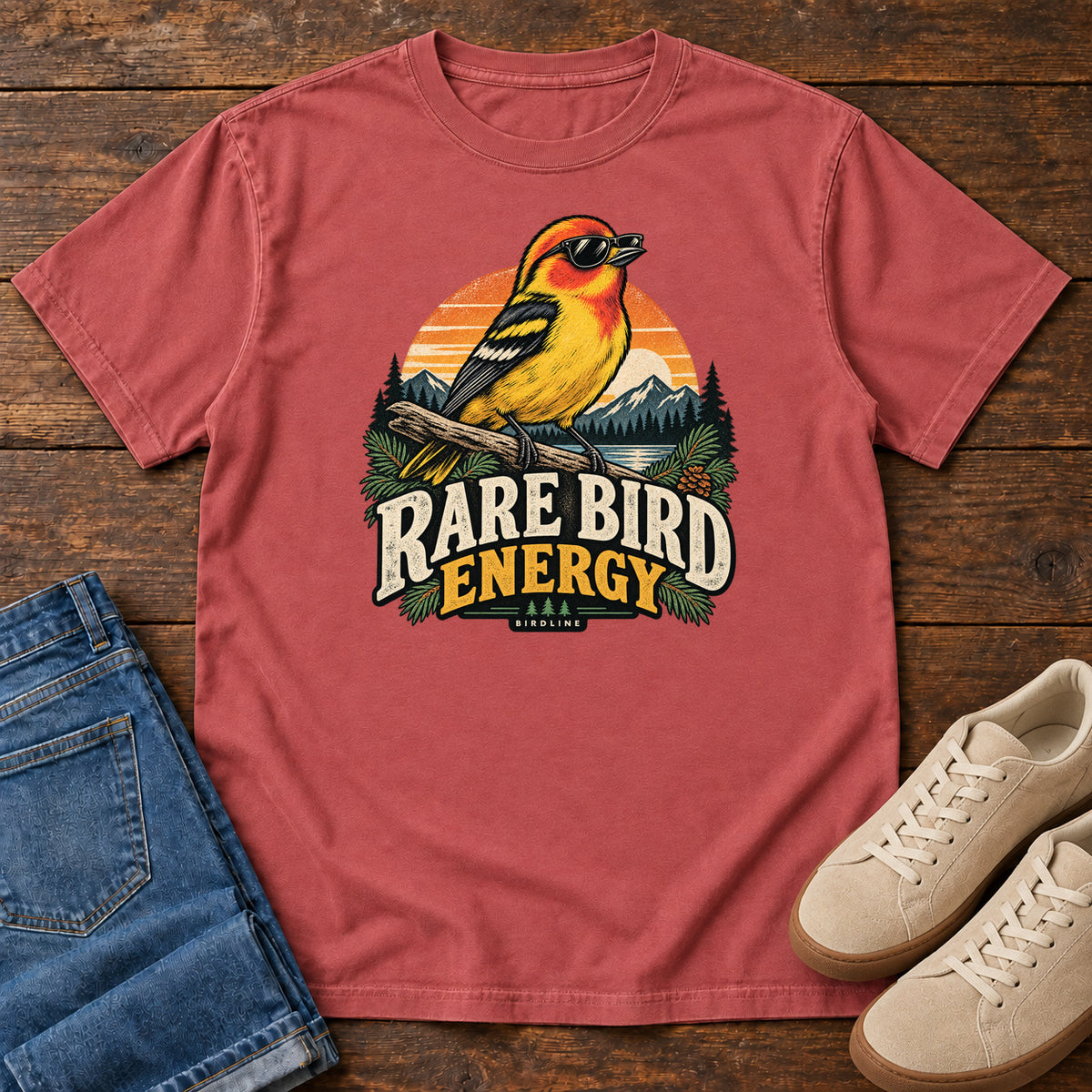 Crimson Rare Bird Energy T-Shirt with a yellow and orange bird wearing sunglasses before a striped sunset; front flat lay on wood.