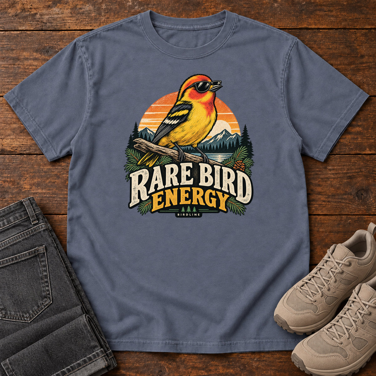 Denim Rare Bird Energy T-Shirt with a yellow and orange bird wearing sunglasses before a striped sunset; front flat lay on wood.