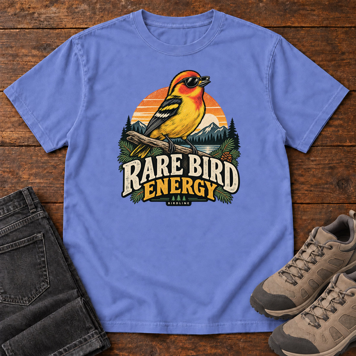 Flo Blue Rare Bird Energy T-Shirt with a yellow and orange bird wearing sunglasses before a striped sunset; front flat lay on wood.