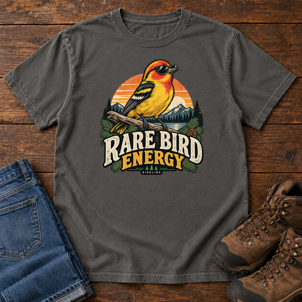 Pepper Rare Bird Energy T-Shirt with a yellow and orange bird wearing sunglasses before a striped sunset; front flat lay on wood.