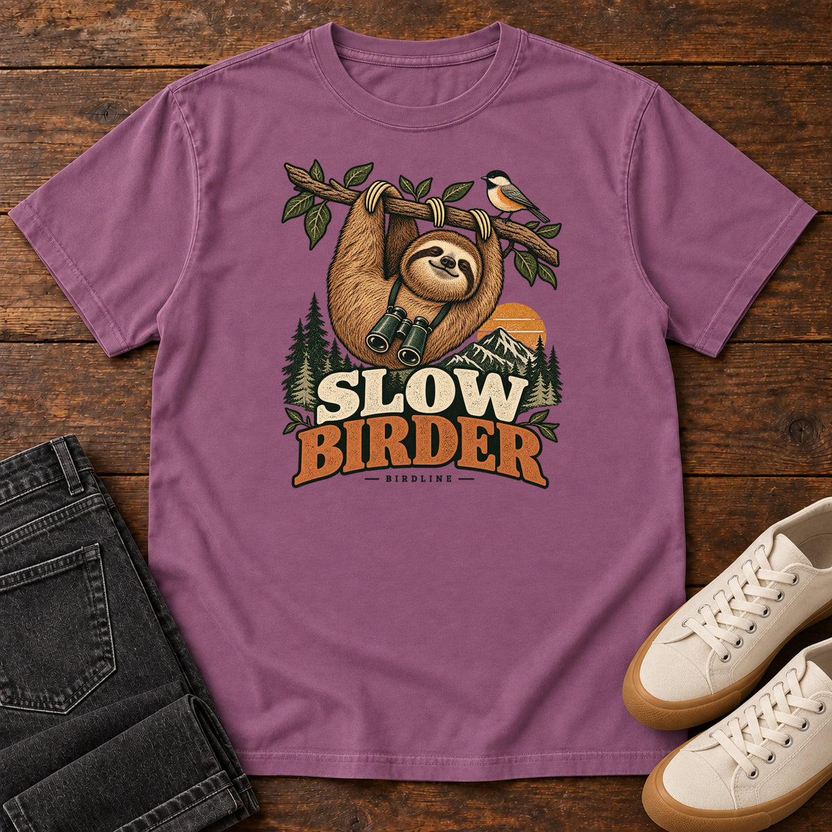 Berry Slow Birder T-Shirt with a sloth with binoculars hanging from a branch beside a bird and mountains; front flat lay on wood.
