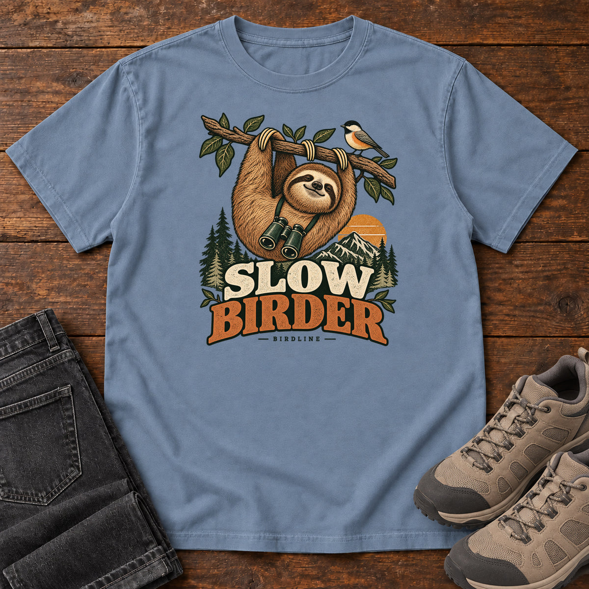 Blue Jean Slow Birder T-Shirt with a sloth with binoculars hanging from a branch beside a bird and mountains; front flat lay on wood.