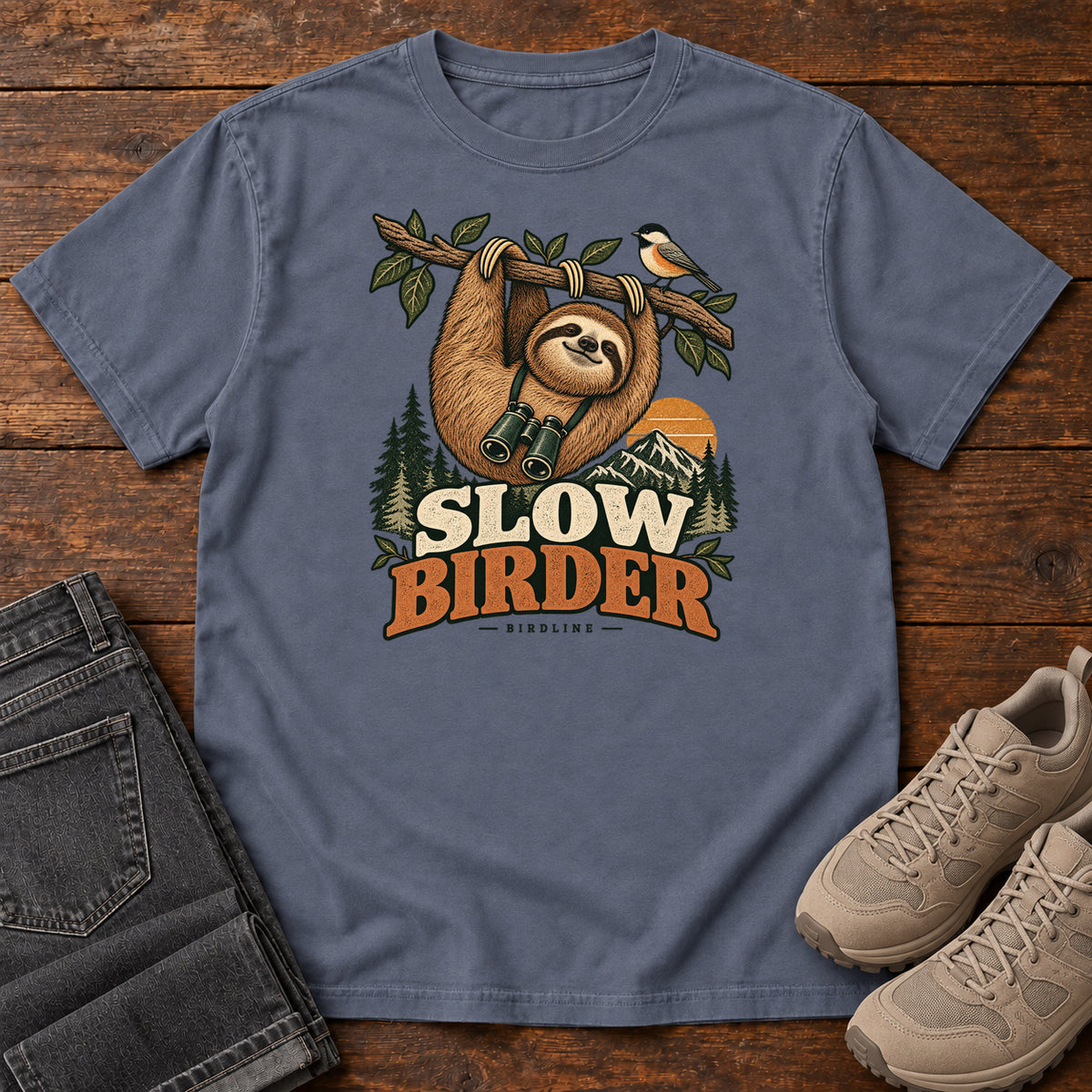 Denim Slow Birder T-Shirt with a sloth with binoculars hanging from a branch beside a bird and mountains; front flat lay on wood.