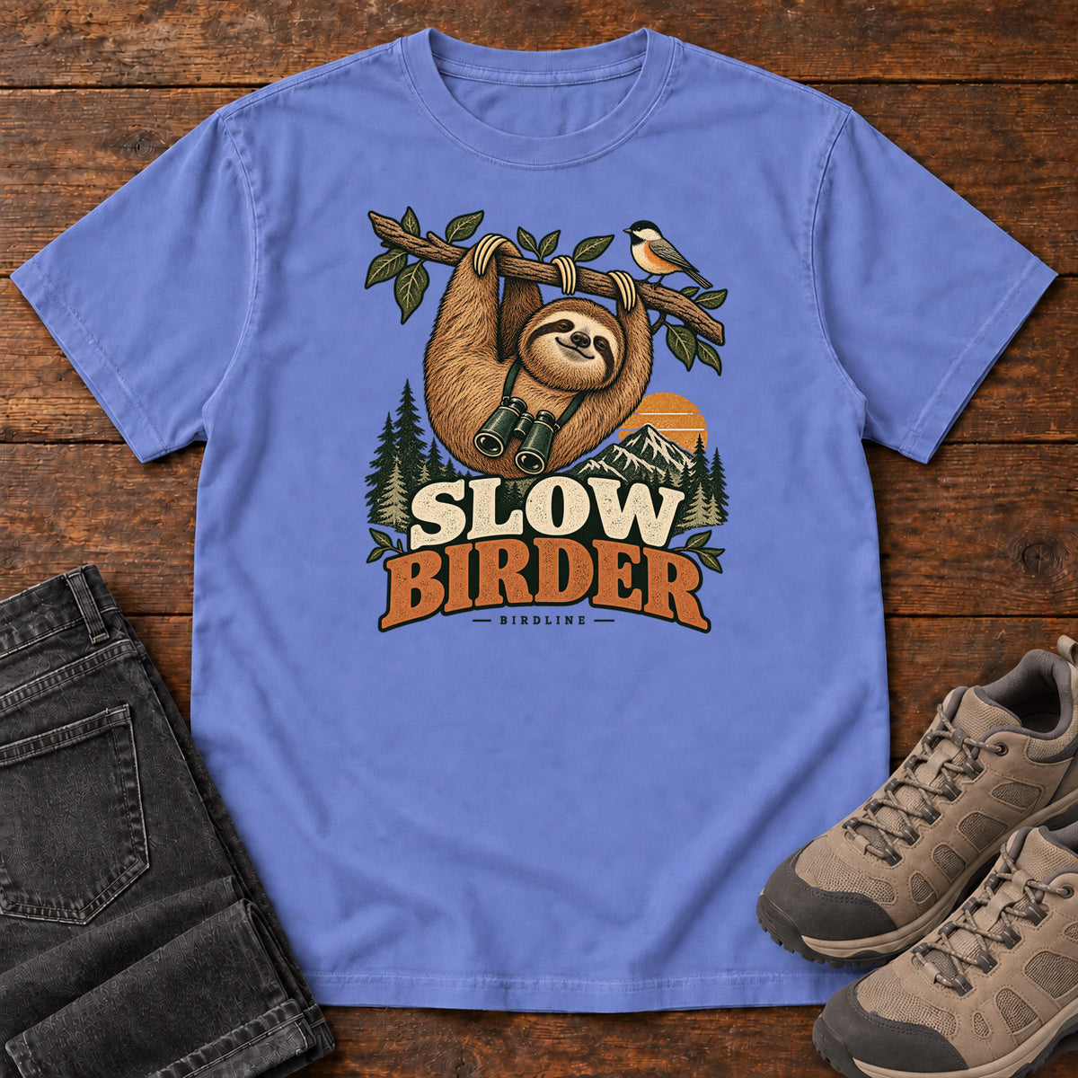 Flo Blue Slow Birder T-Shirt with a sloth with binoculars hanging from a branch beside a bird and mountains; front flat lay on wood.