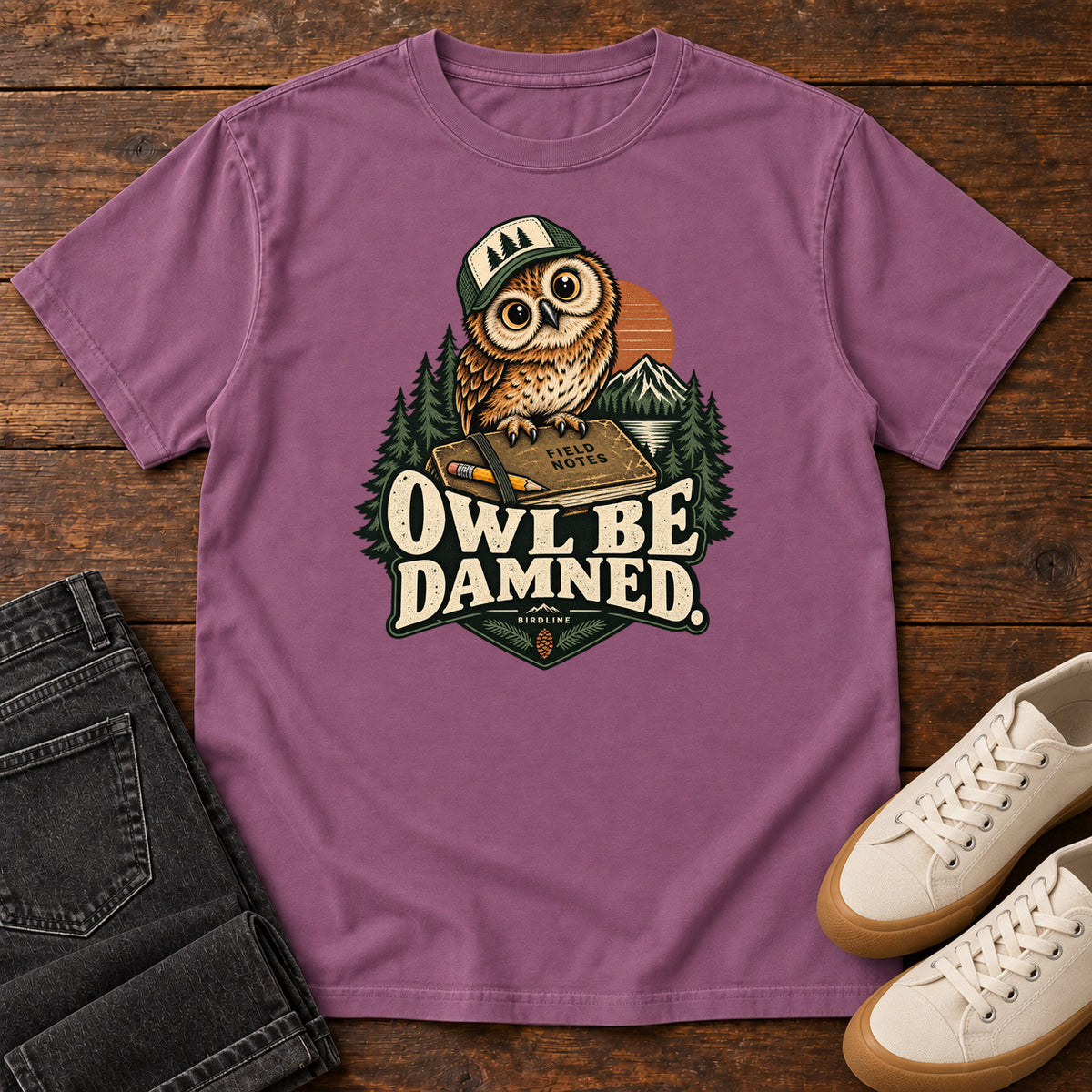 Berry Owl Be Damned. T-Shirt with a brown owl wearing a green cap on a field notebook, with mountains, trees and cream lettering, front flat lay on wood.