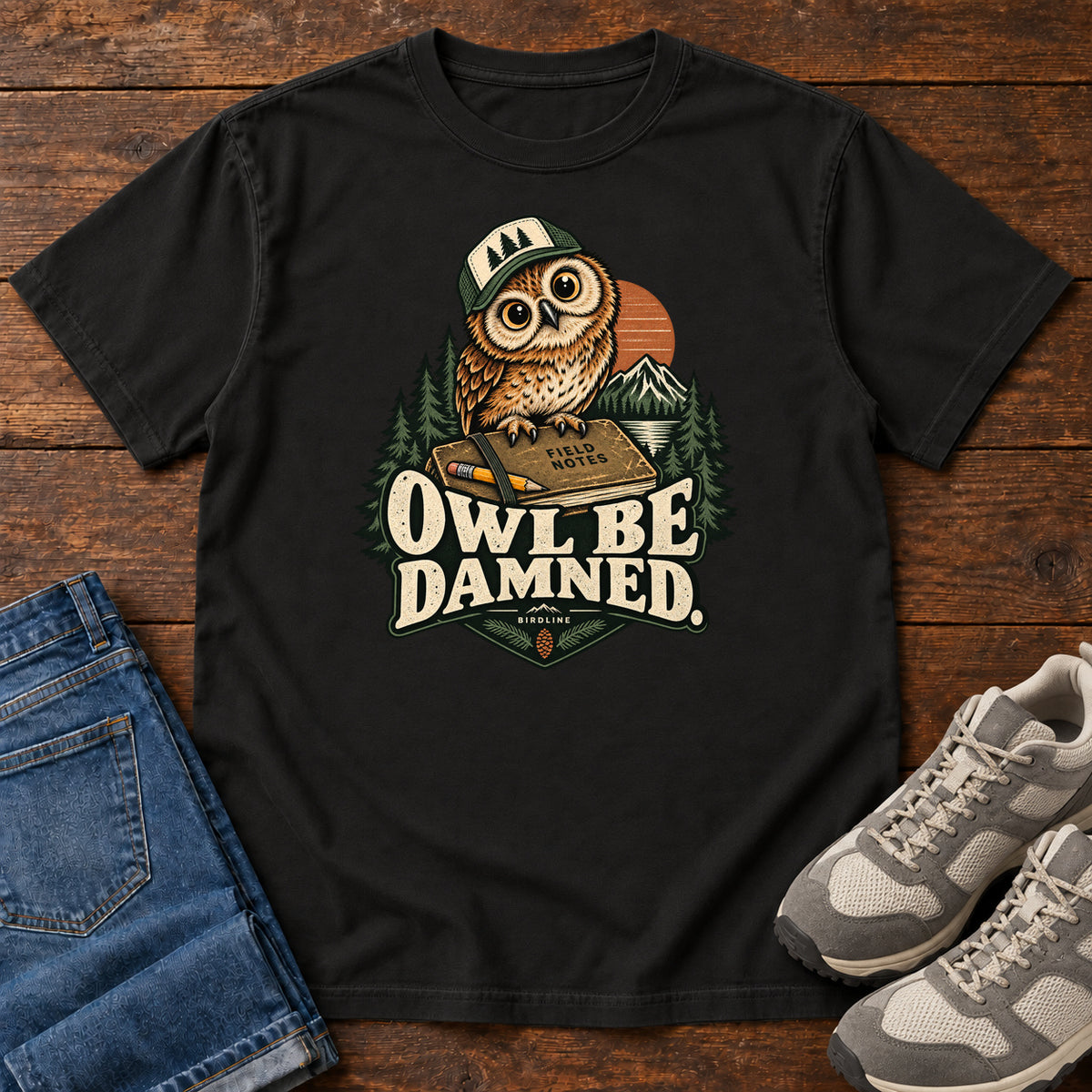 Black Owl Be Damned. T-Shirt with a brown owl wearing a green cap on a field notebook, with mountains, trees and cream lettering, front flat lay on wood.