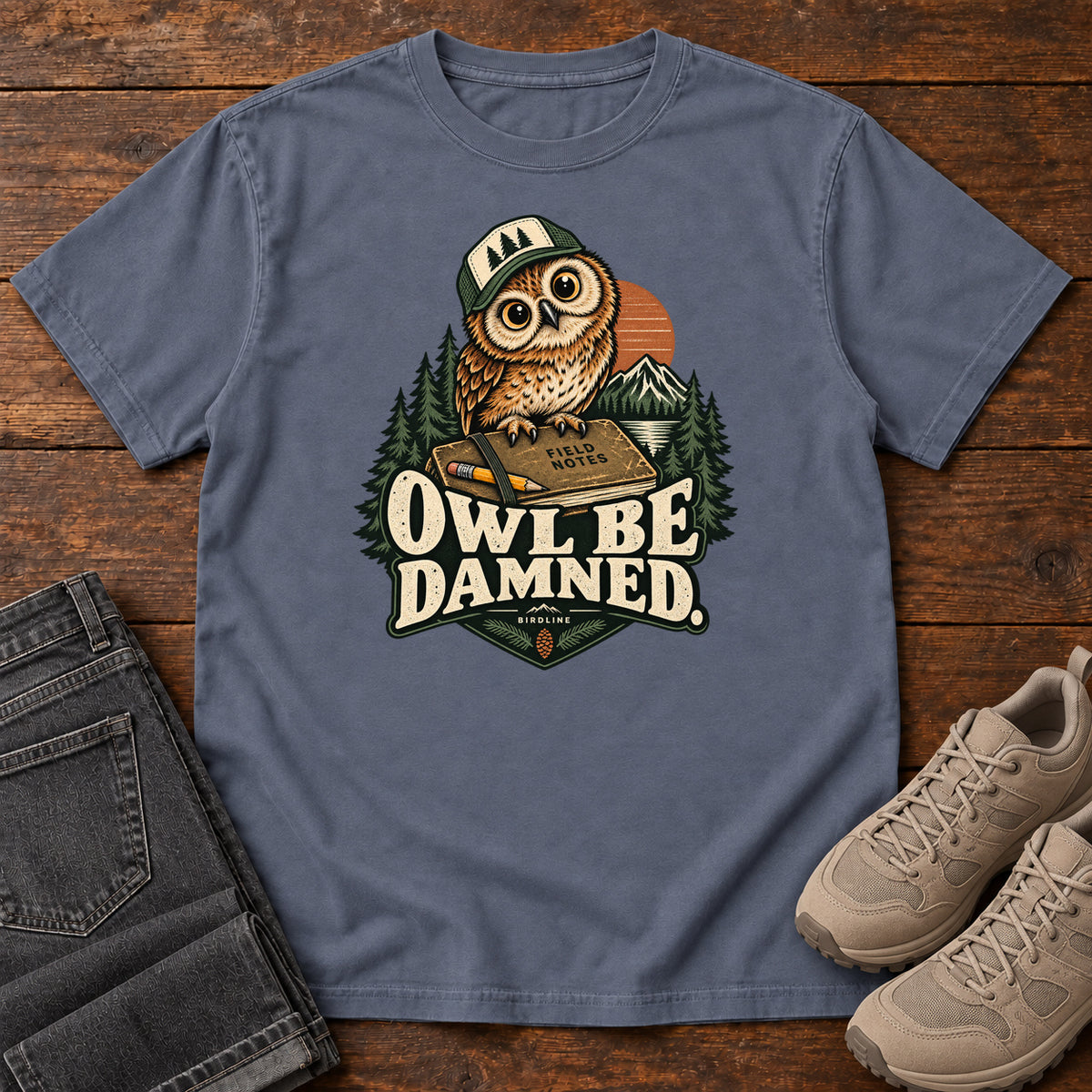 Denim Owl Be Damned. T-Shirt with a brown owl wearing a green cap on a field notebook, with mountains, trees and cream lettering, front flat lay on wood.