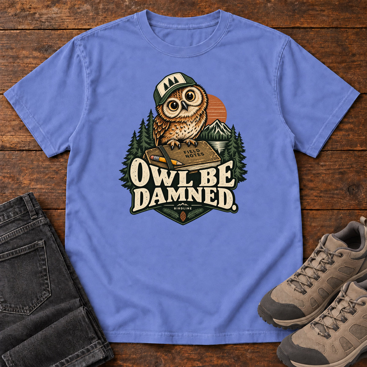 Flo Blue Owl Be Damned. T-Shirt with a brown owl wearing a green cap on a field notebook, with mountains, trees and cream lettering, front flat lay on wood.