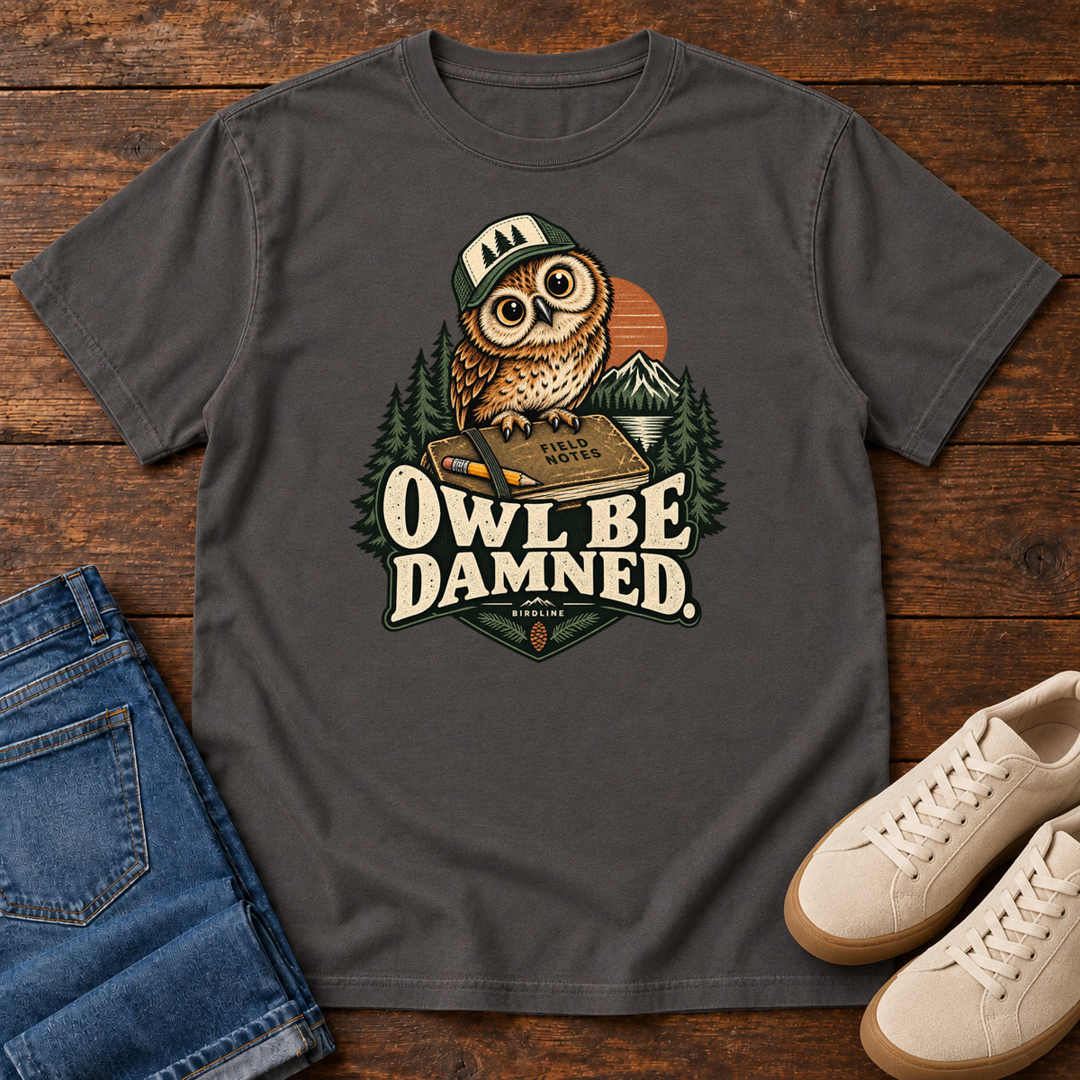 Graphite Owl Be Damned. T-Shirt with a brown owl wearing a green cap on a field notebook, with mountains, trees and cream lettering, front flat lay on wood.
