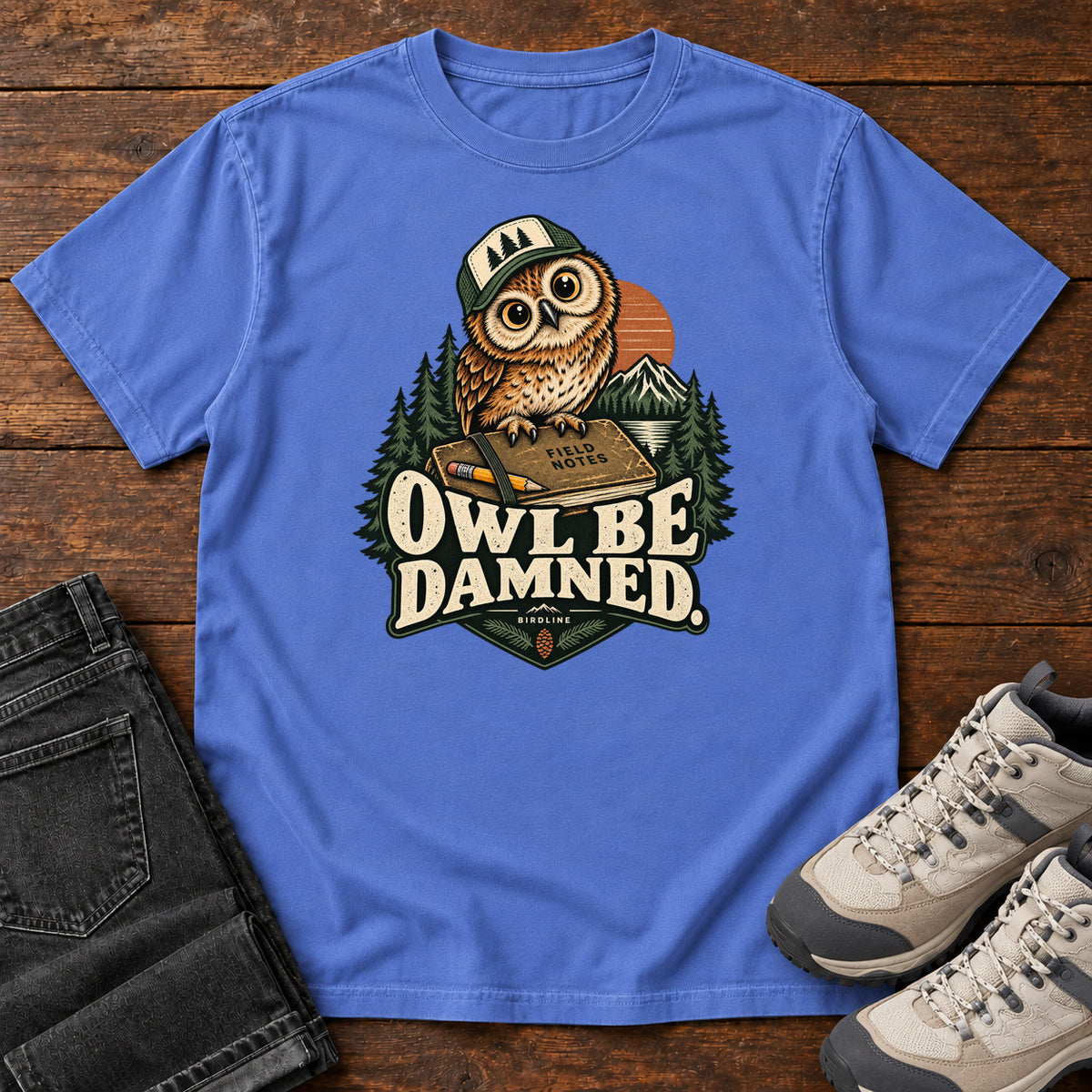 Mystic Blue Owl Be Damned. T-Shirt with a brown owl wearing a green cap on a field notebook, with mountains, trees and cream lettering, front flat lay on wood.