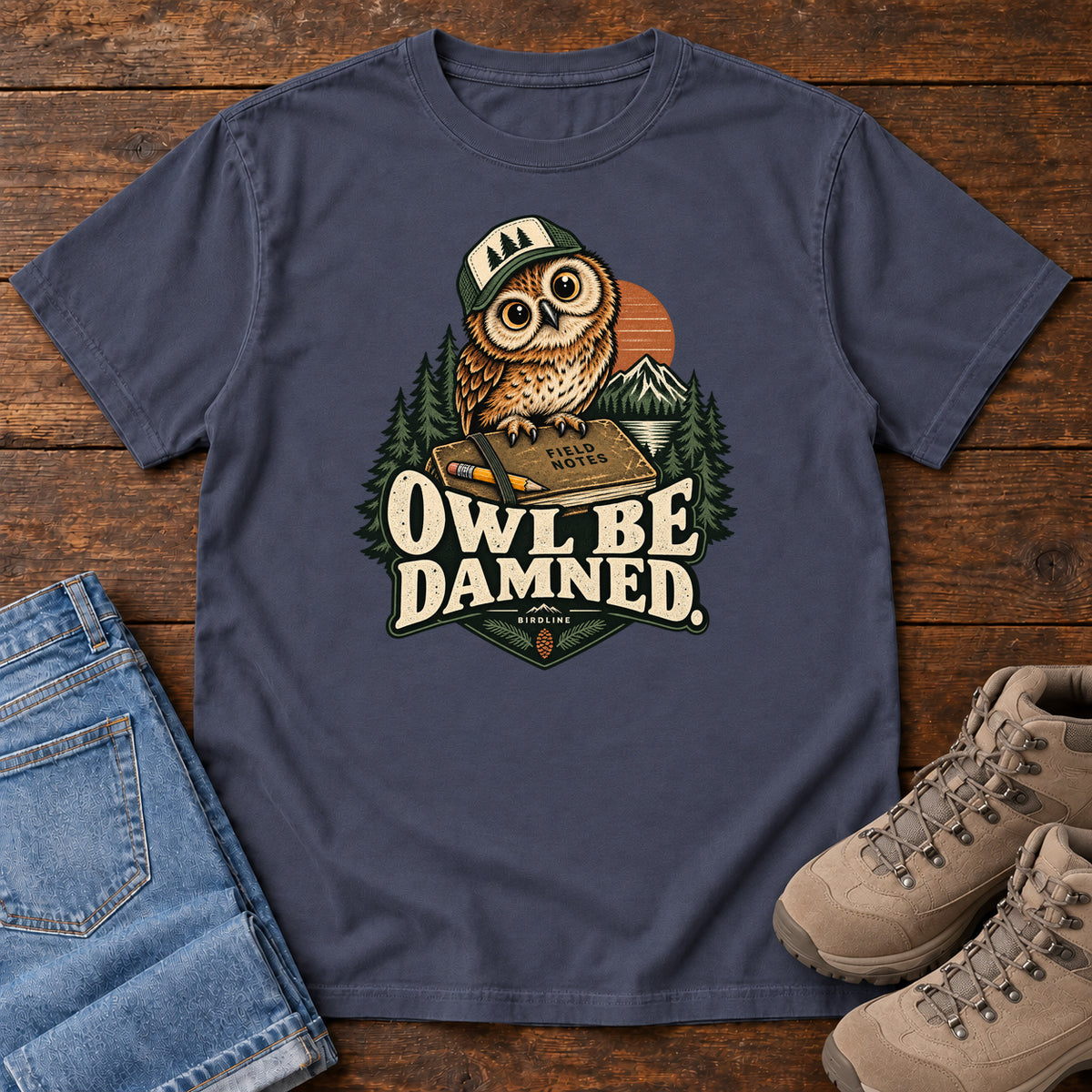 Navy Owl Be Damned. T-Shirt with a brown owl wearing a green cap on a field notebook, with mountains, trees and cream lettering, front flat lay on wood.