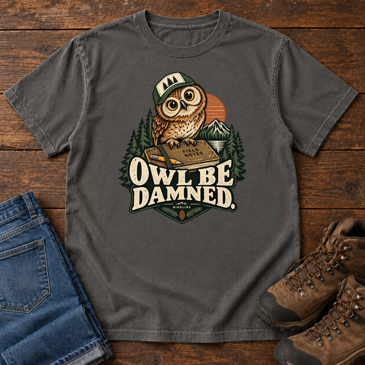 Pepper Owl Be Damned. T-Shirt with a brown owl wearing a green cap on a field notebook, with mountains, trees and cream lettering, front flat lay on wood.