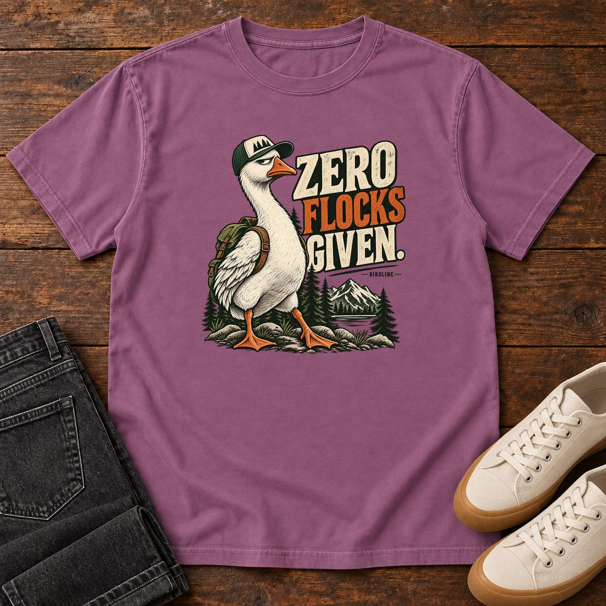 Zero Flocks Given T-Shirt in Berry, front laid flat on wood, featuring a white long-necked bird wearing a cap and backpack beside cream-and-orange lettering and mountains.