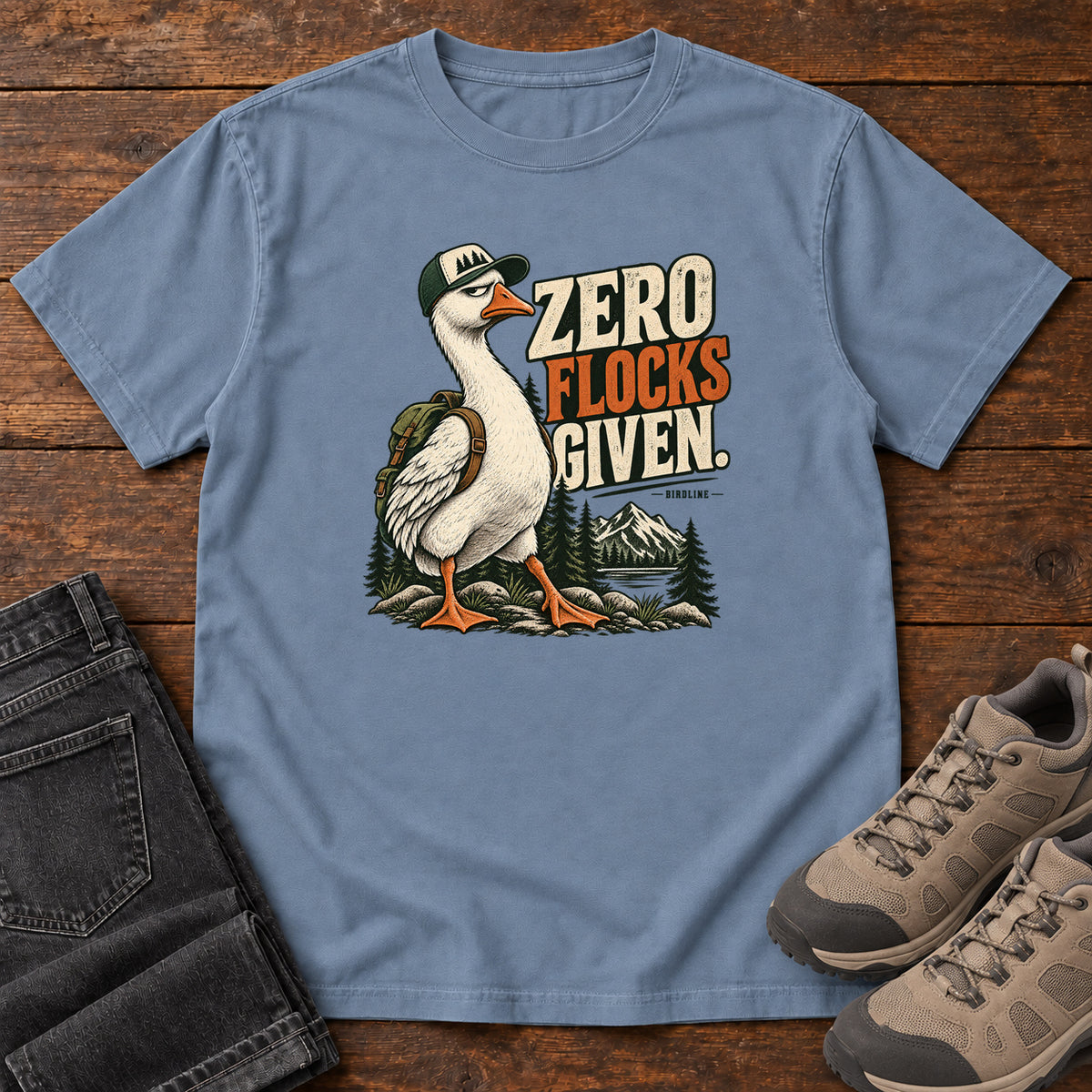 Zero Flocks Given T-Shirt in Blue Jean, front laid flat on wood, featuring a white long-necked bird wearing a cap and backpack beside cream-and-orange lettering and mountains.