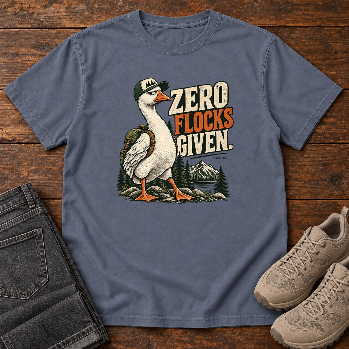 Zero Flocks Given T-Shirt in Denim, front laid flat on wood, featuring a white long-necked bird wearing a cap and backpack beside cream-and-orange lettering and mountains.