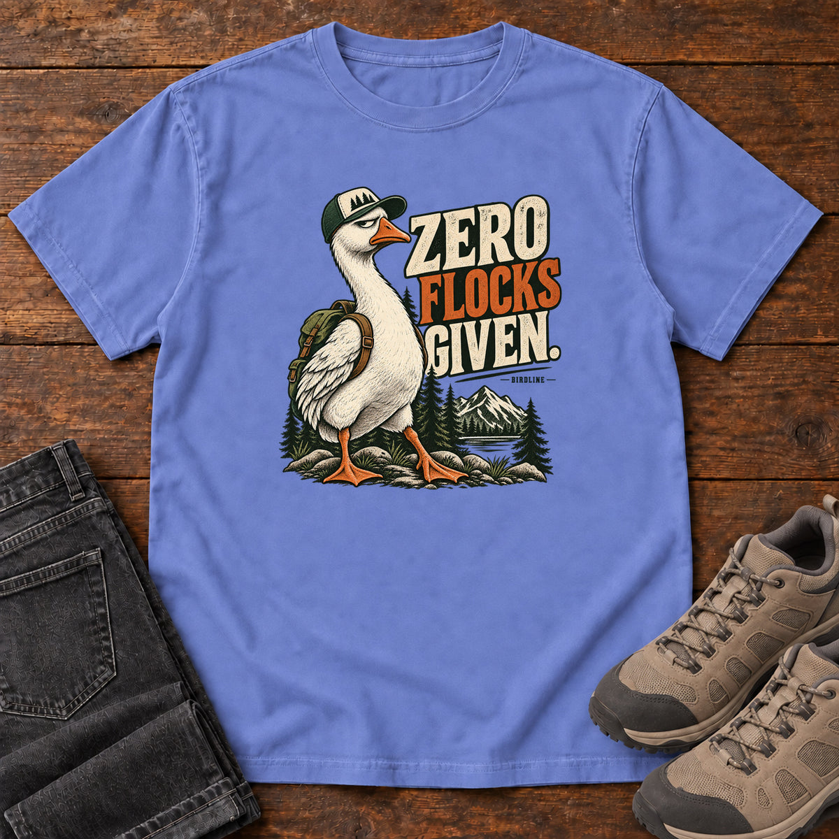 Zero Flocks Given T-Shirt in Flo Blue, front laid flat on wood, featuring a white long-necked bird wearing a cap and backpack beside cream-and-orange lettering and mountains.