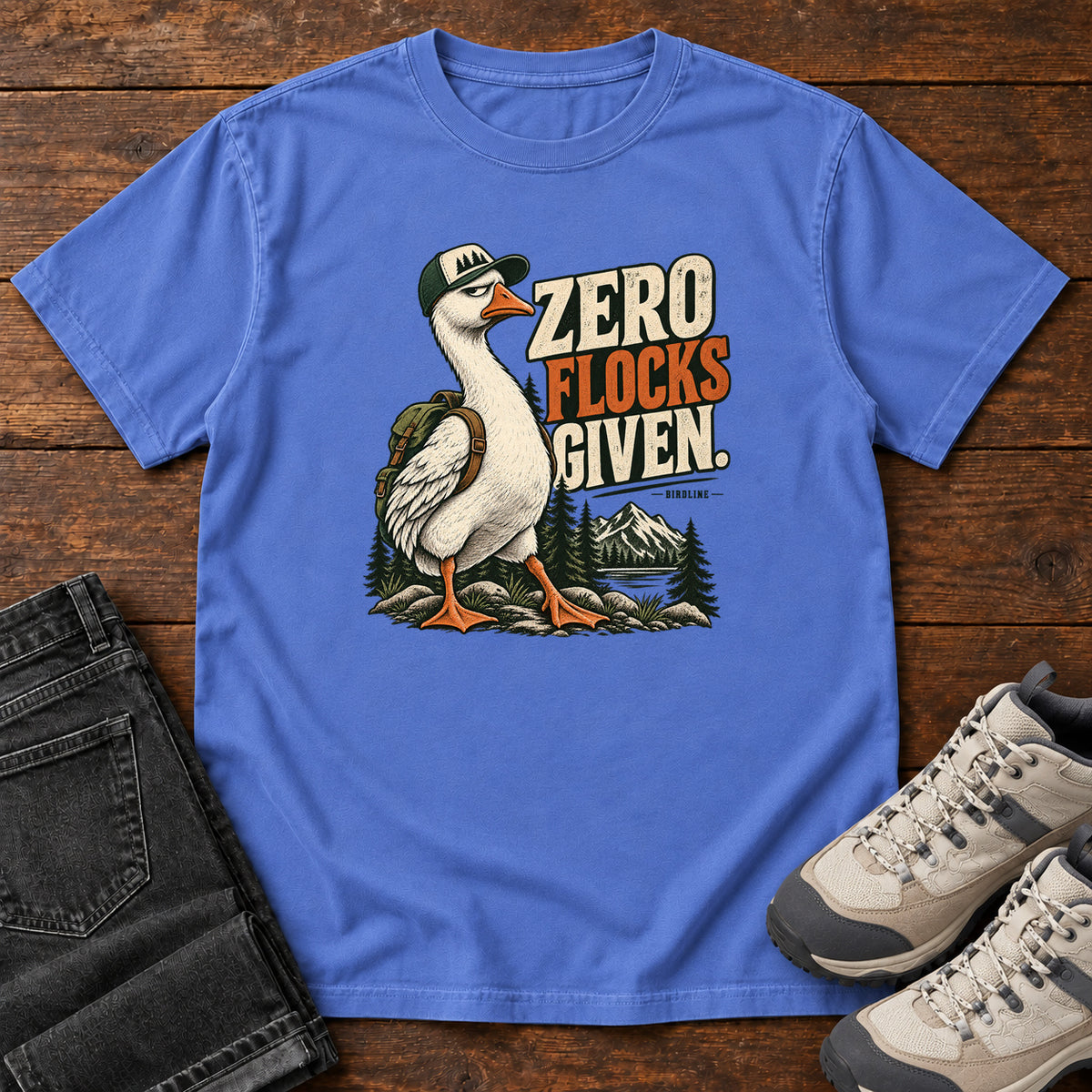 Zero Flocks Given T-Shirt in Mystic Blue, front laid flat on wood, featuring a white long-necked bird wearing a cap and backpack beside cream-and-orange lettering and mountains.