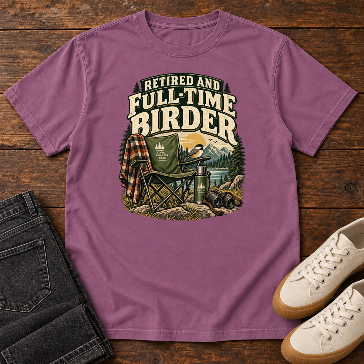 Berry Retired and Full-Time Birder T-Shirt with a camp chair, plaid shirt, flask and binoculars in a mountain-lake scene, front flat lay on wood.