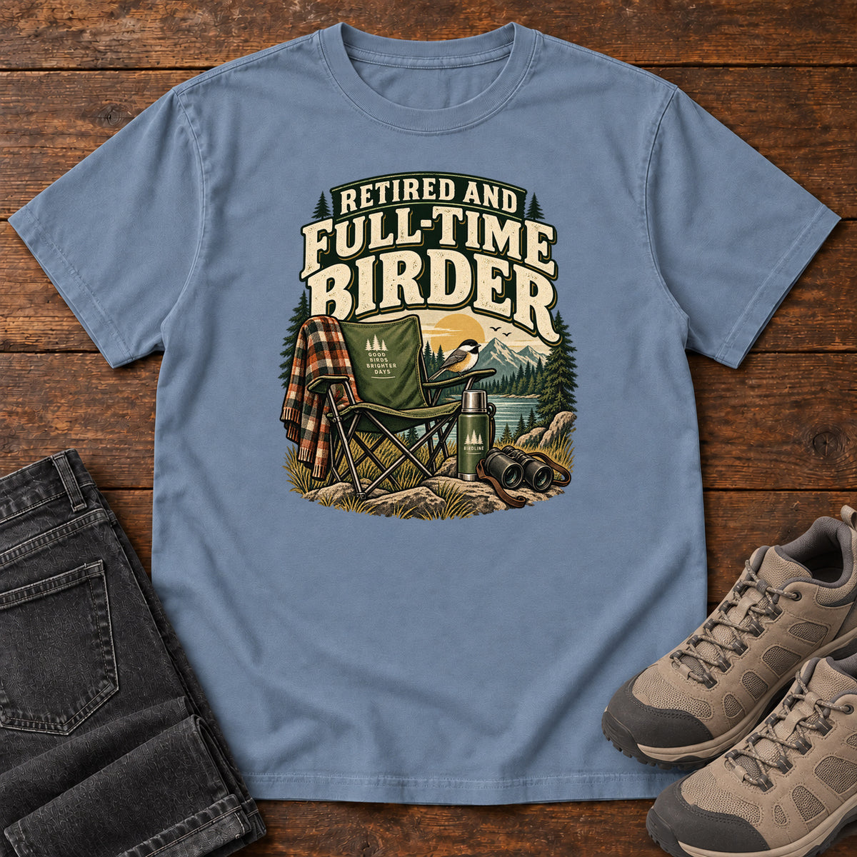 Blue Jean Retired and Full-Time Birder T-Shirt with a camp chair, plaid shirt, flask and binoculars in a mountain-lake scene, front flat lay on wood.