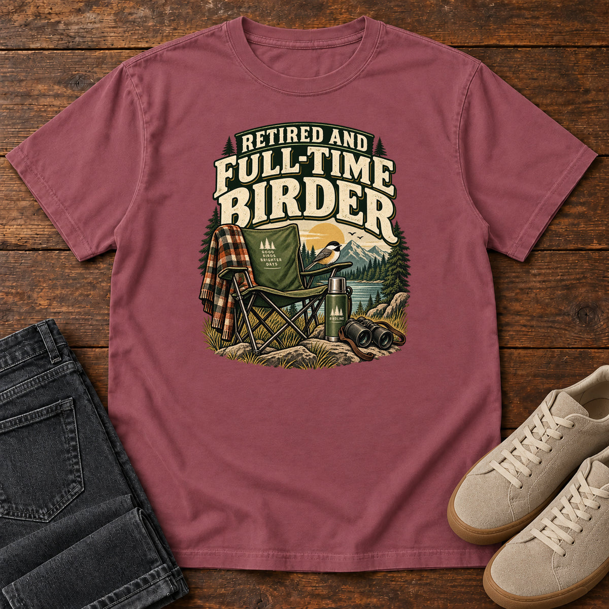 Brick Retired and Full-Time Birder T-Shirt with a camp chair, plaid shirt, flask and binoculars in a mountain-lake scene, front flat lay on wood.