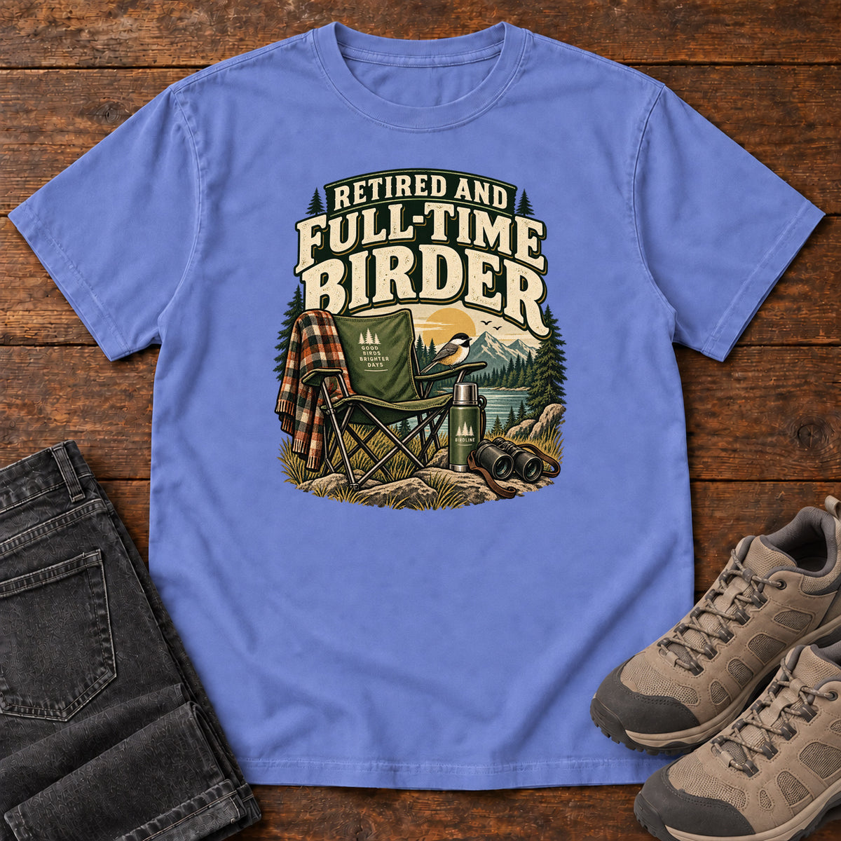 Flo Blue Retired and Full-Time Birder T-Shirt with a camp chair, plaid shirt, flask and binoculars in a mountain-lake scene, front flat lay on wood.
