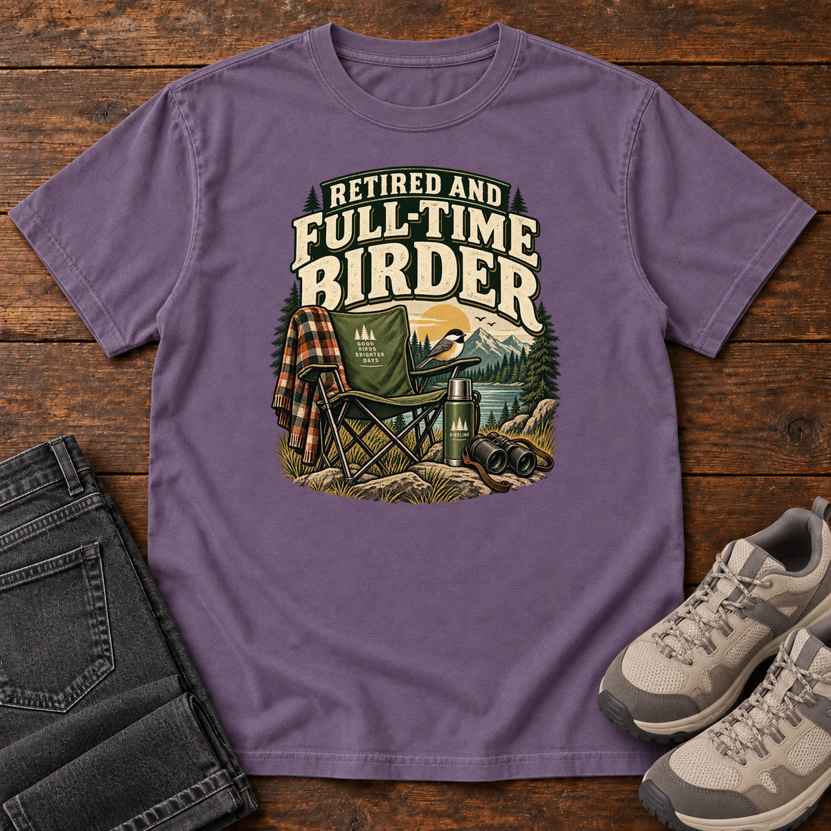 Grape Retired and Full-Time Birder T-Shirt with a camp chair, plaid shirt, flask and binoculars in a mountain-lake scene, front flat lay on wood.