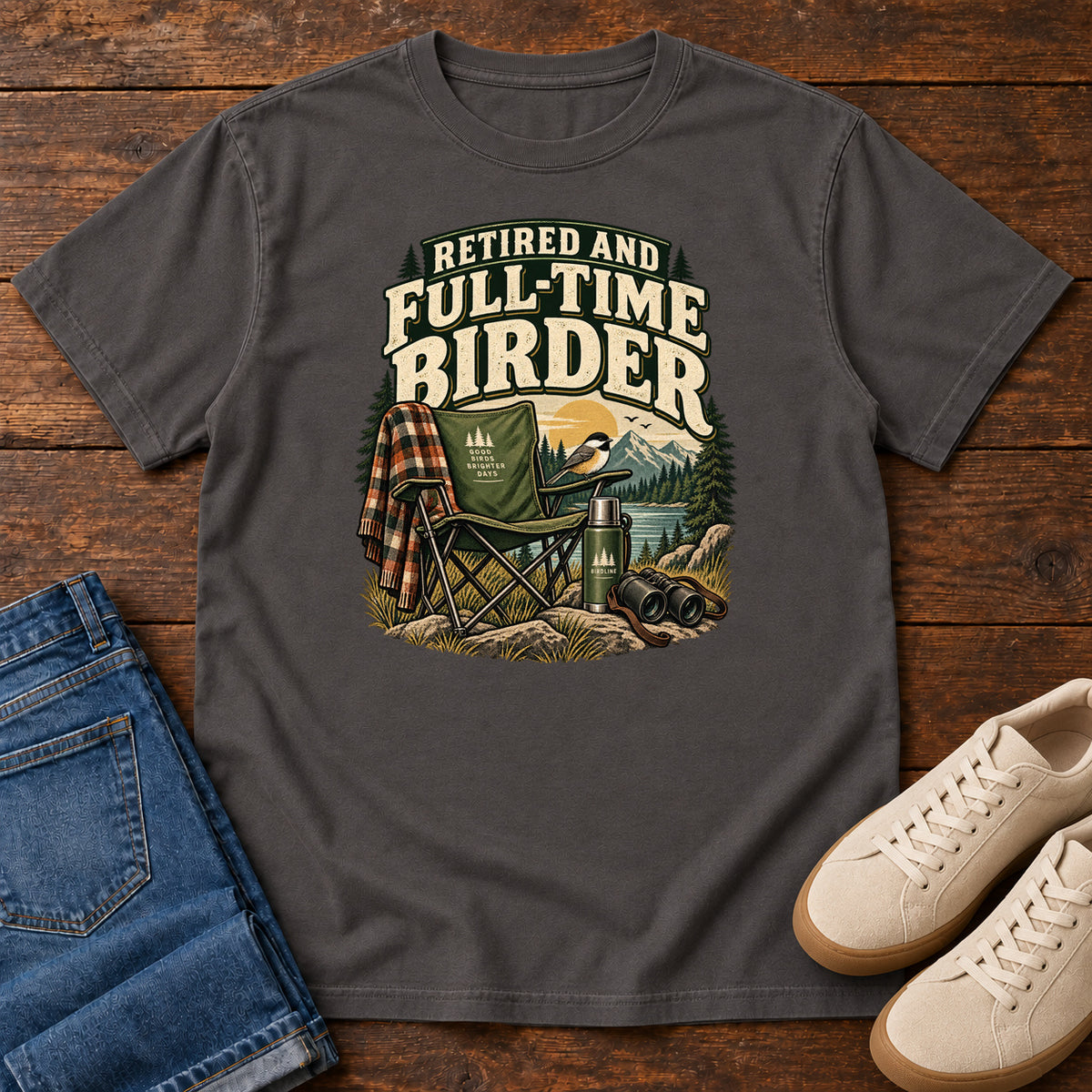 Graphite Retired and Full-Time Birder T-Shirt with a camp chair, plaid shirt, flask and binoculars in a mountain-lake scene, front flat lay on wood.