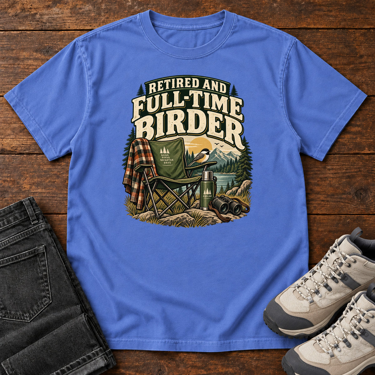 Mystic Blue Retired and Full-Time Birder T-Shirt with a camp chair, plaid shirt, flask and binoculars in a mountain-lake scene, front flat lay on wood.