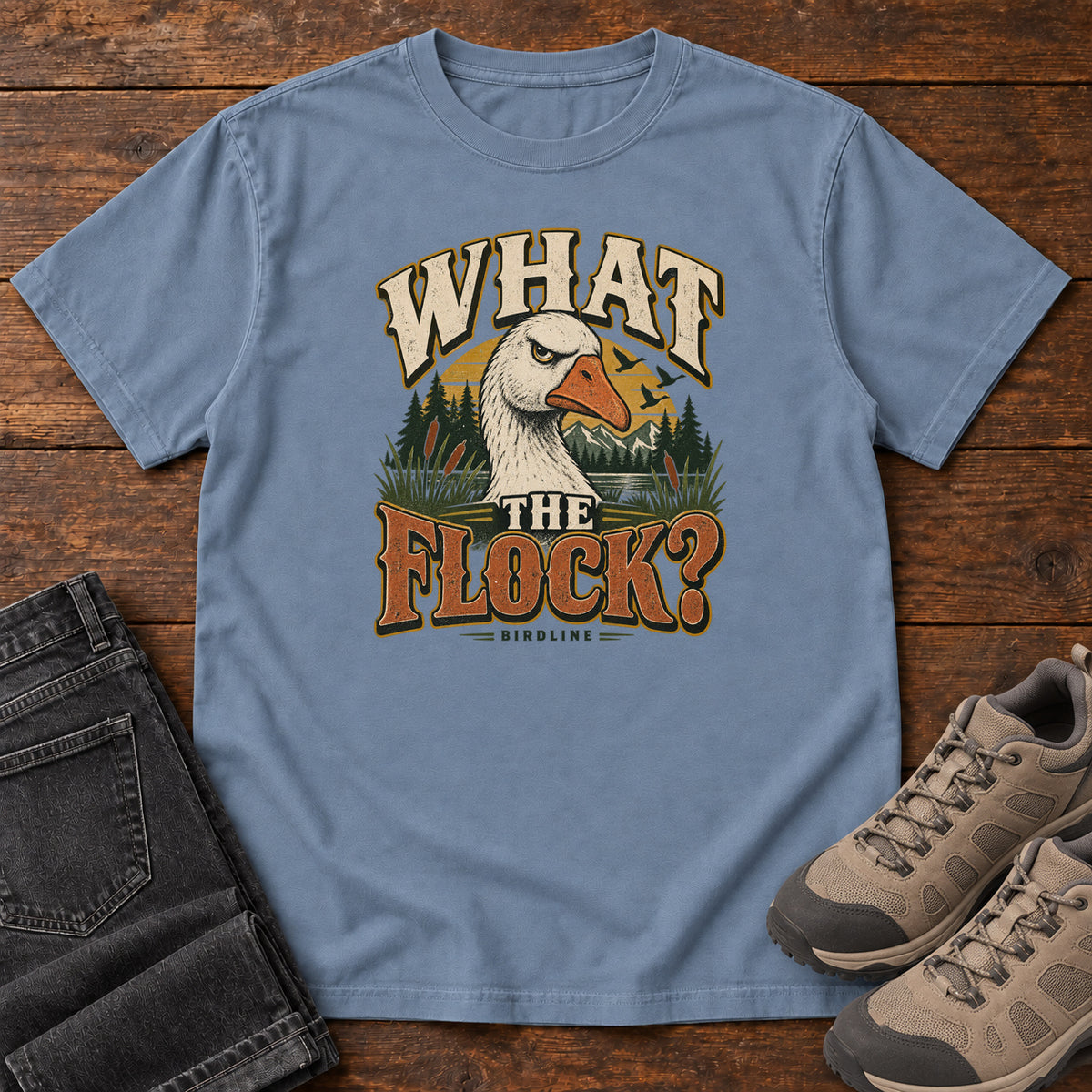 Blue Jean What the Flock? T-Shirt with a white goose head, evergreens and cream and orange slogan lettering; front flat lay on wood.