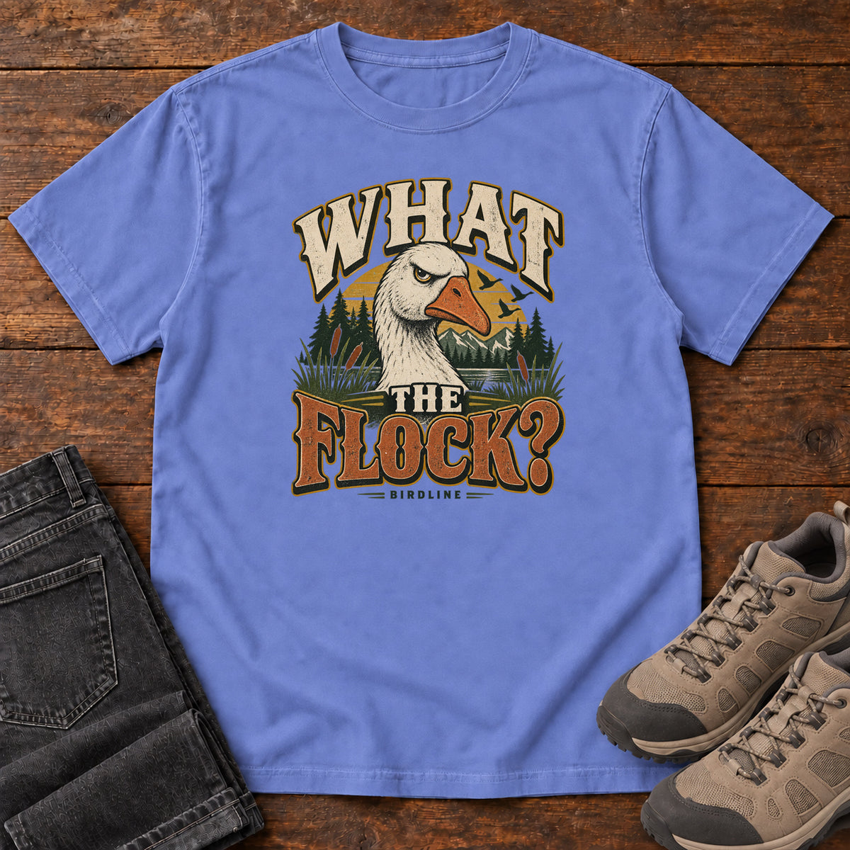 Flo Blue What the Flock? T-Shirt with a white goose head, evergreens and cream and orange slogan lettering; front flat lay on wood.