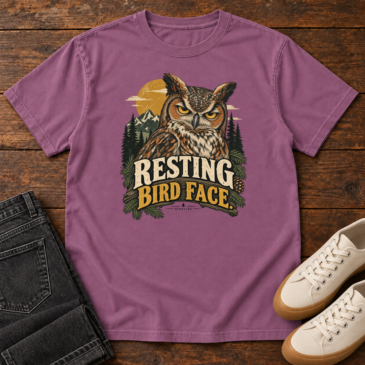 Berry Resting Bird Face T-Shirt with a yellow-eyed, tufted owl with pine branches, mountains and a golden sun; front flat lay on wood.