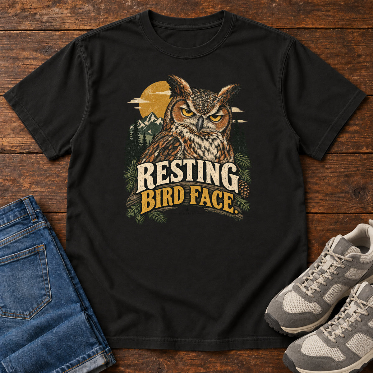 Black Resting Bird Face T-Shirt with a yellow-eyed, tufted owl with pine branches, mountains and a golden sun; front flat lay on wood.