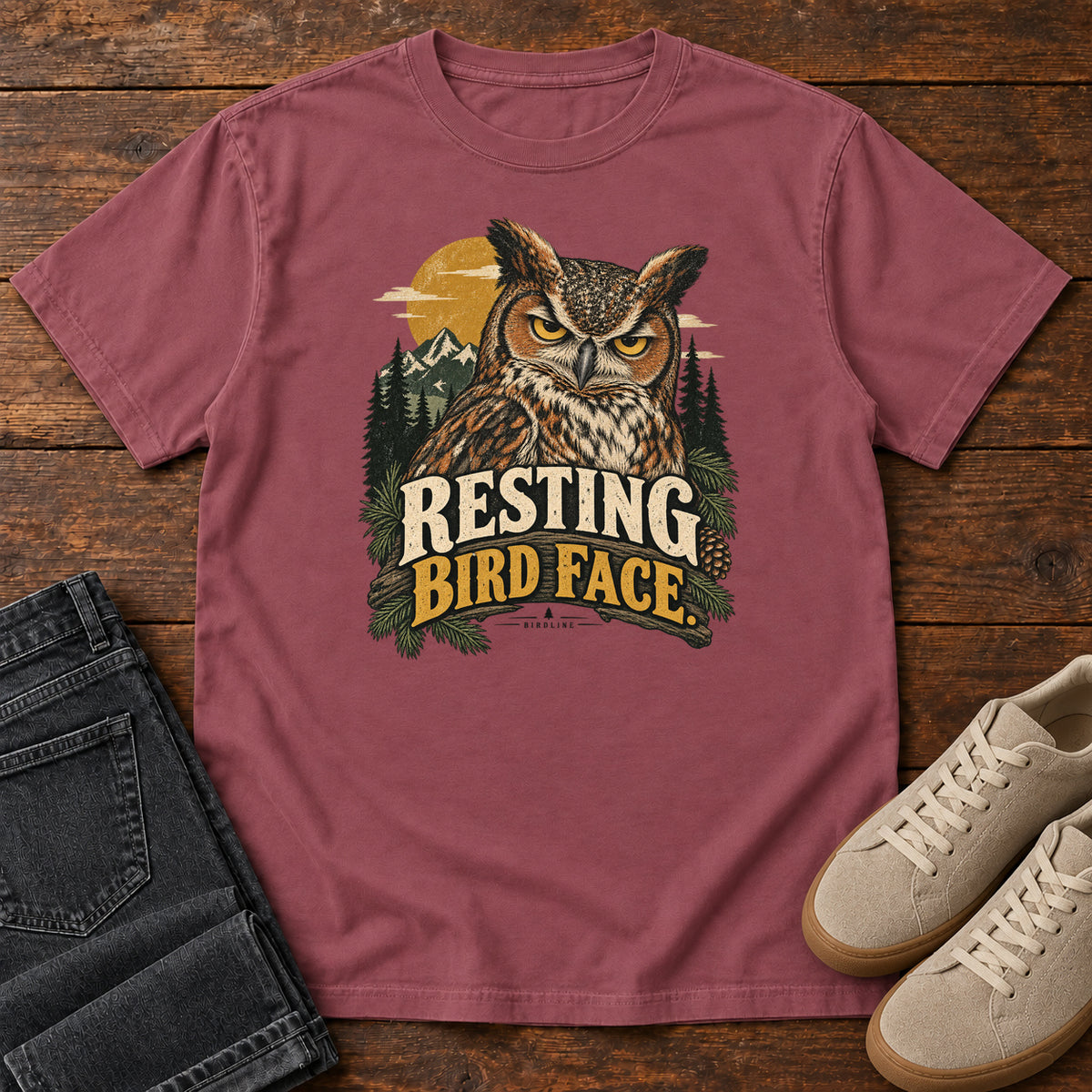 Brick Resting Bird Face T-Shirt with a yellow-eyed, tufted owl with pine branches, mountains and a golden sun; front flat lay on wood.