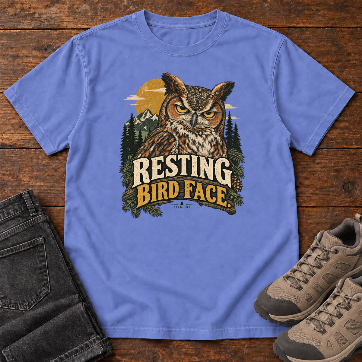 Flo Blue Resting Bird Face T-Shirt with a yellow-eyed, tufted owl with pine branches, mountains and a golden sun; front flat lay on wood.
