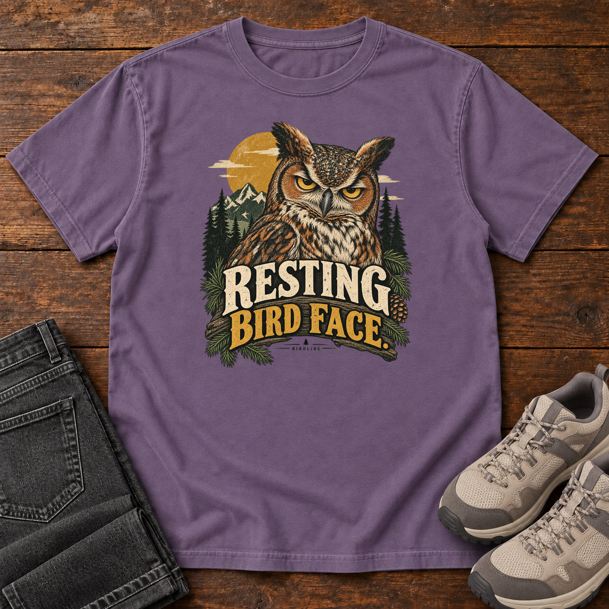 Grape Resting Bird Face T-Shirt with a yellow-eyed, tufted owl with pine branches, mountains and a golden sun; front flat lay on wood.