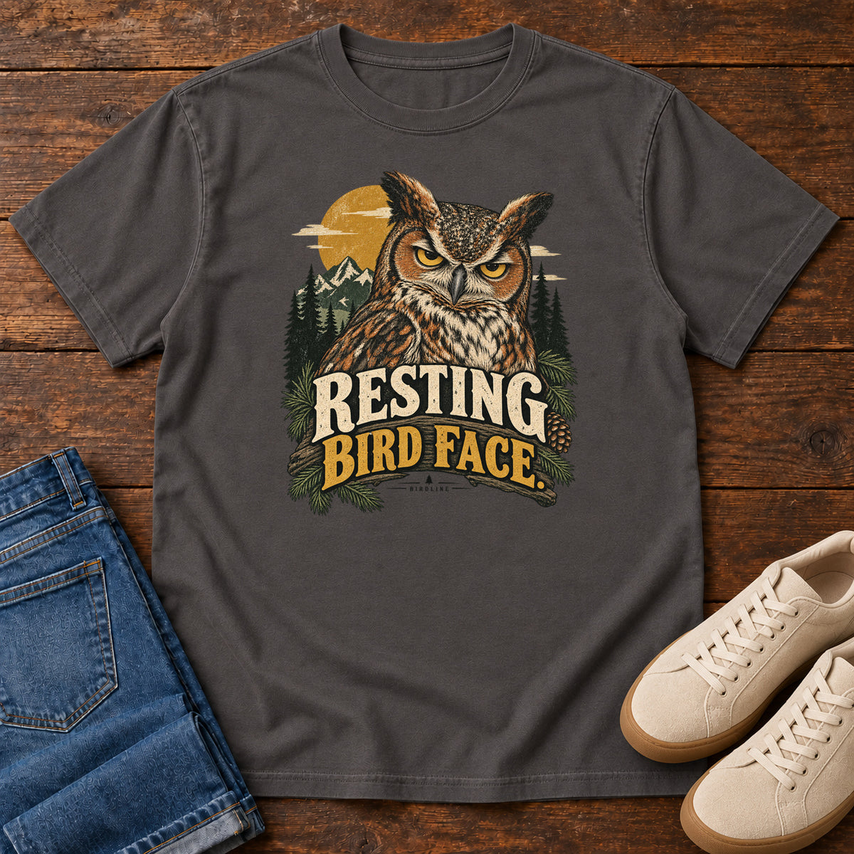 Graphite Resting Bird Face T-Shirt with a yellow-eyed, tufted owl with pine branches, mountains and a golden sun; front flat lay on wood.