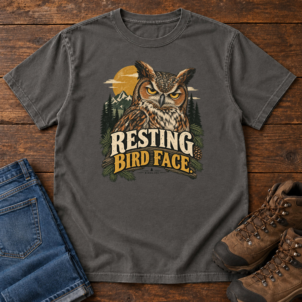Pepper Resting Bird Face T-Shirt with a yellow-eyed, tufted owl with pine branches, mountains and a golden sun; front flat lay on wood.